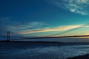 Silhouette of a suspension bridge against a twilight sky with clouds and a calm river in the foreground. photo