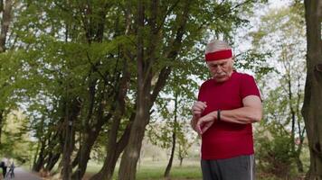 Senior corredor homem dentro parque usando inteligente assistir, rastreamento distância, verificação pulso depois de ginástica exercite-se video