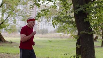 mayor antiguo hombre correr, trabajando fuera cardio en parque y utilizando Bluetooth auriculares, escuchando música video