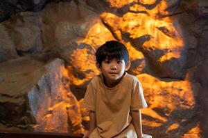 un pequeño asiático chico se sienta en contra un antecedentes de rocas, naranja luces salpicaduras el antecedentes. foto