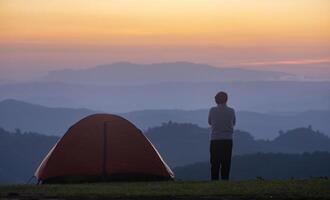 Traveller is standing by the tent during overnight camping while looking at the beautiful scenic sunset over the mountain for outdoor adventure vacation travel concept photo