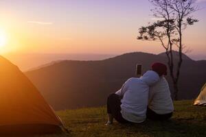 Couple is sitting by the tent during overnight camping and taking selfie while looking at the beautiful scenic sunset over the mountain for outdoor adventure vacation travel photo