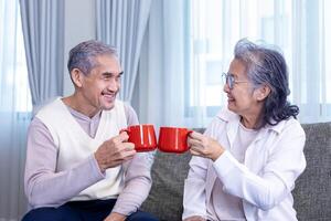 Pareja de sano asiático mayor padre y madre sentado en sofá con contento sonrisa a Jubilación hogar Bebiendo caliente té a celebrar su fiesta juntos para mayor cuidado y gasto valioso hora foto