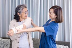 Senior Asian woman got medical service visit from caregiver nurse at home while doing physical therapy to shoulder stretching for muscle pain in health care and pension welfare concept photo