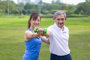 Senior asian man is using dumbbell for weight training strength while his daughter is supporting in the public park for elder longevity exercise and outdoor workout usage photo