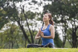 joven asiático mujer en yoga traje relajantemente practicando meditación en el bosque a alcanzar felicidad desde interior paz sabiduría para respiración ejercicio, sano mente y alma foto