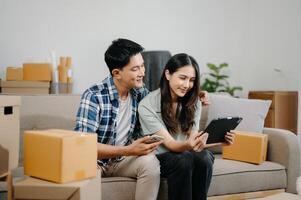 joven atractivo Pareja hombre y mujer utilizar tableta y teléfono inteligente en línea compras mueble Decorar casa con caja de cartón paquete moverse en nuevo casa. foto