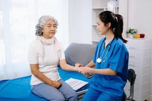médico en blanco traje tomar notas mientras que se discute y asiático anciano, mujer paciente quien acostado en cama con recepción salina solución foto