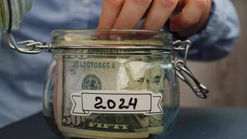 femmina mani prendere i soldi a partire dal bicchiere vaso pieno di americano moneta dollari denaro contante banconota con testo 2024. preparazione Salvataggio i soldi. moderare consumo e economia. raccolta i soldi video