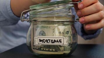 Unrecognizable woman add money to Saving Money In Glass Jar filled with Dollars banknotes. MORTGAGE transcription in front of jar. Managing personal finances extra income for future insecurity background video