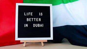 Frame text LIFE IS BETTER IN DUBAI on background of waving UAE flag made from silk. United Arab Emirates flag with concept of tourism and traveling. Inviting greeting card, advertisement. Dubai welcoming card video