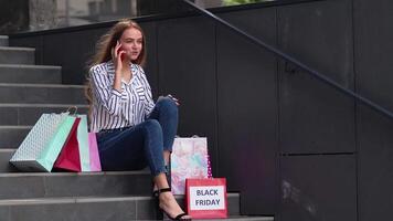 menina sentado em escadas com bolsas falando em Móvel telefone sobre venda dentro compras Shopping dentro Preto Sexta-feira video