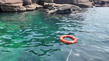 Orange lifebuoy floating on calm turquoise sea water with rocky coastline in the background, representing safety and rescue concepts video