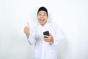 stunned face of asian muslim man showing thumbs up with holding phone looking at camera photo