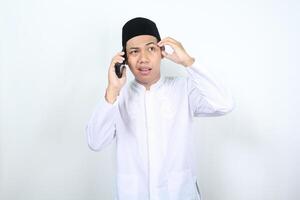 asiático musulmán hombre conmovedor su cabeza espectáculo confuso expresión mientras hablando en el teléfono aislado en blanco antecedentes foto