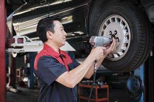 One expert Asian male automotive mechanic technician is screwing car wheel nuts on lifting with drill wrench for repair at garage. Vehicle maintenance service works industry occupation business jobs. photo