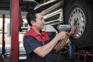 One expert Asian male automotive mechanic technician is screwing car wheel nuts on lifting with drill wrench for repair at garage. Vehicle maintenance service works industry occupation business jobs. photo