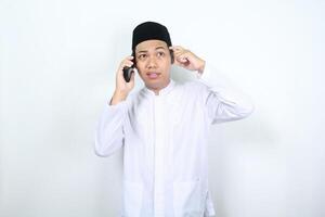 asiático musulmán hombre conmovedor su cabeza espectáculo confuso expresión mientras hablando en el teléfono aislado en blanco antecedentes foto