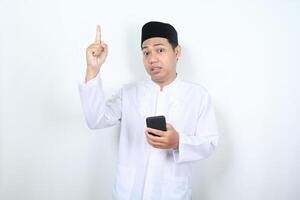 asian muslim man holding mobile phone pointing above with confused expression isolated on white background photo