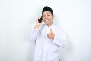 smiling muslim asian man showing thumbs up while calling with his mobile phone isolated photo