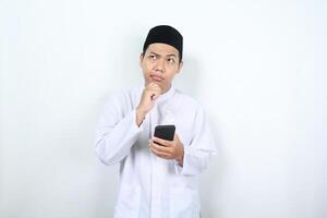 thoughtful asian muslim man looking above with hand on chin thinking an offer while holding mobile phone isolated on white background photo