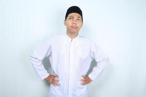 thoughtful asian muslim man looking up with hand on waist thinking something isolated on white background photo