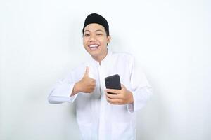 sonriente asiático musulmán hombre participación teléfono y demostración pulgares arriba aislado foto