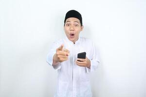 amazed asian muslim man pointing finger to camera with holding smartphone isolated photo