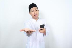 asian muslim man holding mobile phone presenting hand forward to camera with confused expression isolated on white background photo