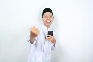 successful asian muslim man raising fist to celebrate victory with holding phone isolated photo