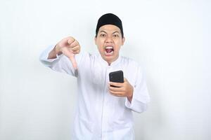 enojado asiático musulmán hombre gritando con demostración pulgar abajo mientras participación móvil teléfono foto