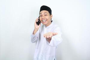 delighted asian muslim man presenting hand forward to camera with smiling face while speaking on his phone isolated photo