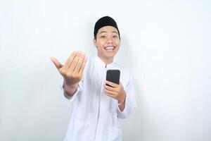 smiling asian muslim man showing come on gesture with holding phone isolated photo