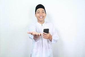 pleased asian muslim man smiling while presenting hand forward to camera with holding mobile phone isolated photo