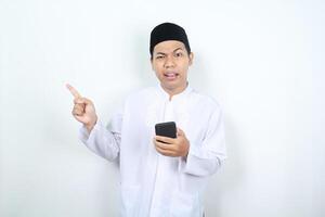 asian muslim man holding mobile phone pointing to beside with confused expression isolated on white background photo