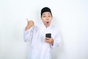 shocked asian muslim man holding phone and pointing to the right side isolated photo