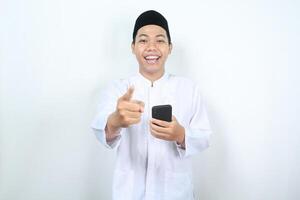 happy asian muslim man smiling while pointing hand to camera with holding mobile phone isolated photo