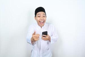 excited muslim asian man pointing hand to camera with holding phone photo