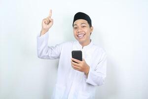 smiling asian muslim man pointing to above with holding mobile phone isolated photo