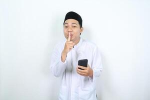 smiling asian muslim man holding phone show silent gesture with finger on lips isolated photo