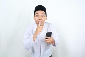 smiling asian muslim man holding phone show silent gesture with finger on lips isolated photo