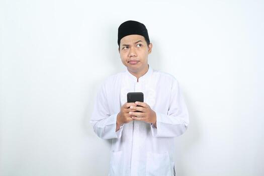 confused muslim asian man looking above think about something with holding mobile phone isolated on white background photo
