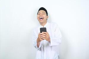 cheerful asian muslim man laughing while holding phone isolated photo