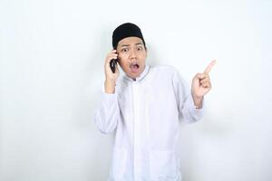 shocked asian muslim man pointing to beside while talking on mobile phone photo