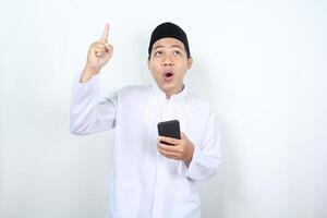 asiático musulmán hombre conmocionado mirando encima con señalando dedo arriba y participación móvil teléfono aislado foto