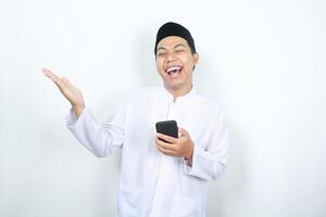 joyful asian muslim man laughing while holding phone with presenting hand to the right side isolated photo