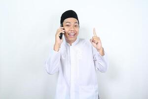 cheerful muslim asian man pointing to above while speaking on his phone with smiling face isolated photo
