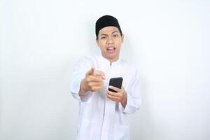 asian muslim man holding mobile phone pointing forward at camera with confused expression isolated on white background photo
