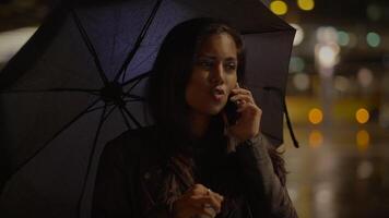 jovem mulher falando em Móvel telefone lado de fora dentro a chuva às noite video