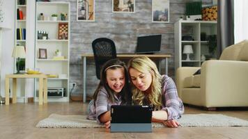 alegre pequeño niña con su madre acostado en el piso utilizando tableta computadora. video